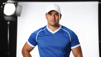 A 30-year-old coach with a strong build, wearing a blue jersey with white stripes, and a white hat, standing in front of...