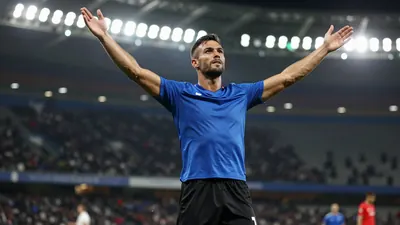 Photorealistic image of a muscular male footballer in a solid blue jersey and black shorts, celebrating a goal with arms...