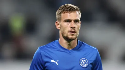 A mid-30s male Vålerenga player in a solid blue kit, head-and-shoulders, looking determined, background of a blurred...